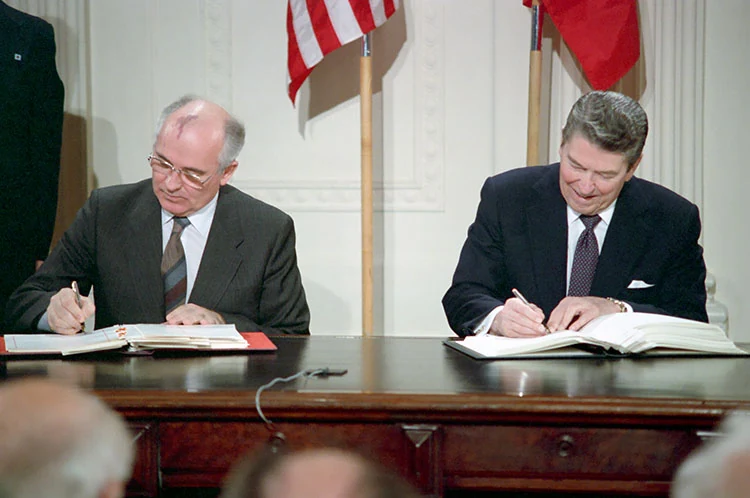 Mikhail Gorbachev and Ronald Reagan sit beside one another signing treaty documents in the White House East Room.