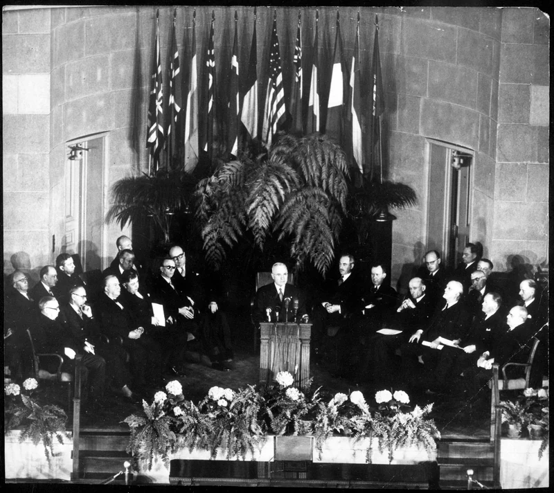 Harry S. Truman speaks at a podium with the foreign ministers of the NATO signatory nations seated behind him.