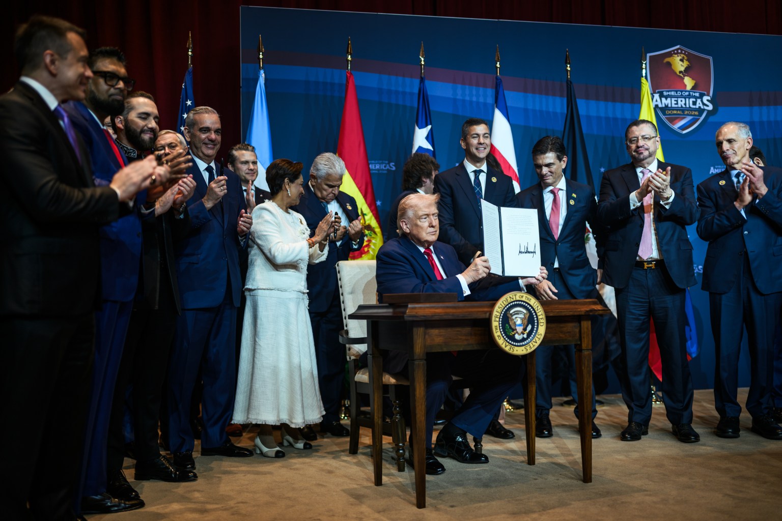 Donald Trump holds an open document folder at a table while other summit participants stand behind him.